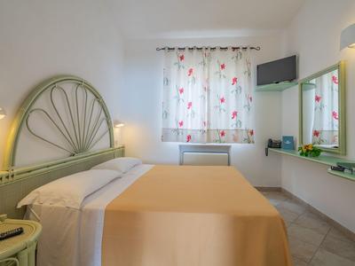 Ein schlicht eingerichtetes Hotelzimmer mit Doppelbett, grünem Kopfteil und Blumenvorhang am Fenster.