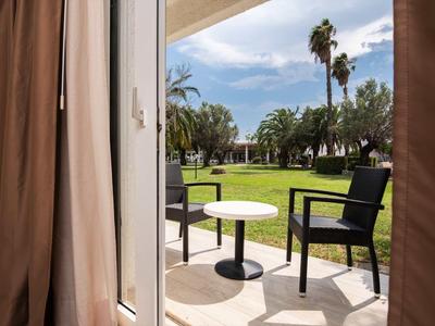 Widok z pokoju z brązowymi zasłonami na taras z dwoma krzesłami i małym stolikiem, dalej trawnik i palmy.