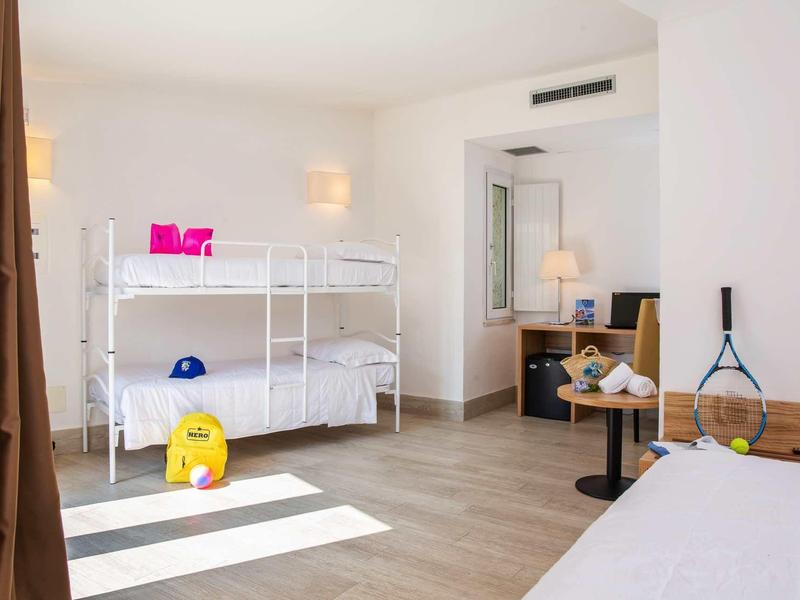 Jasno umeblowany pokój hotelowy z łóżkiem piętrowym, biurkiem i kolorowymi zabawkami dla dzieci na podłodze.