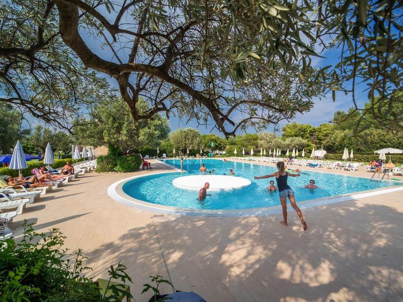 Okrągły basen na zewnątrz z leżakami i parasolami pod zielonymi drzewami.