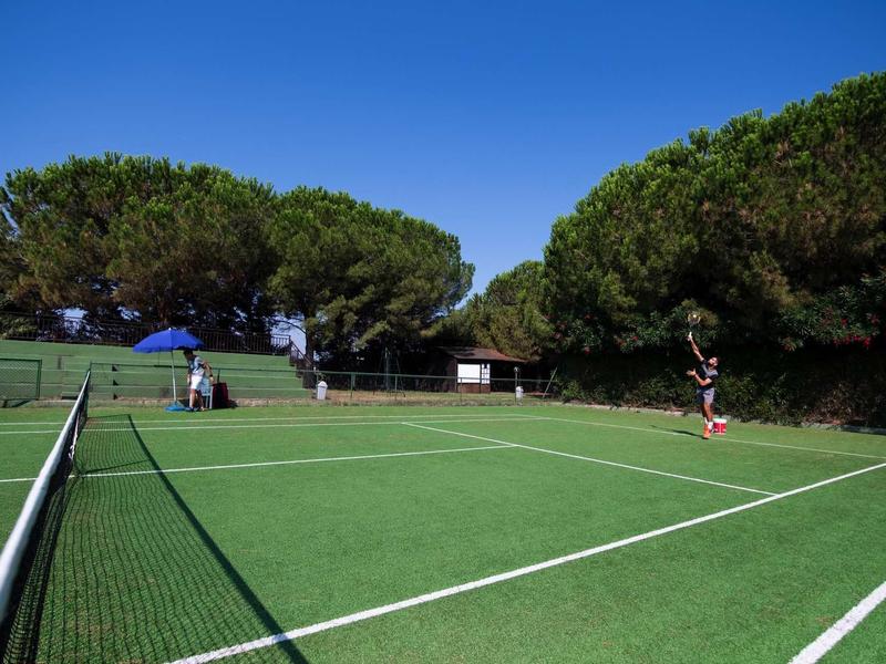 Kort tenisowy na świeżym powietrzu z graczami, otoczony drzewami i niebieskim niebem.