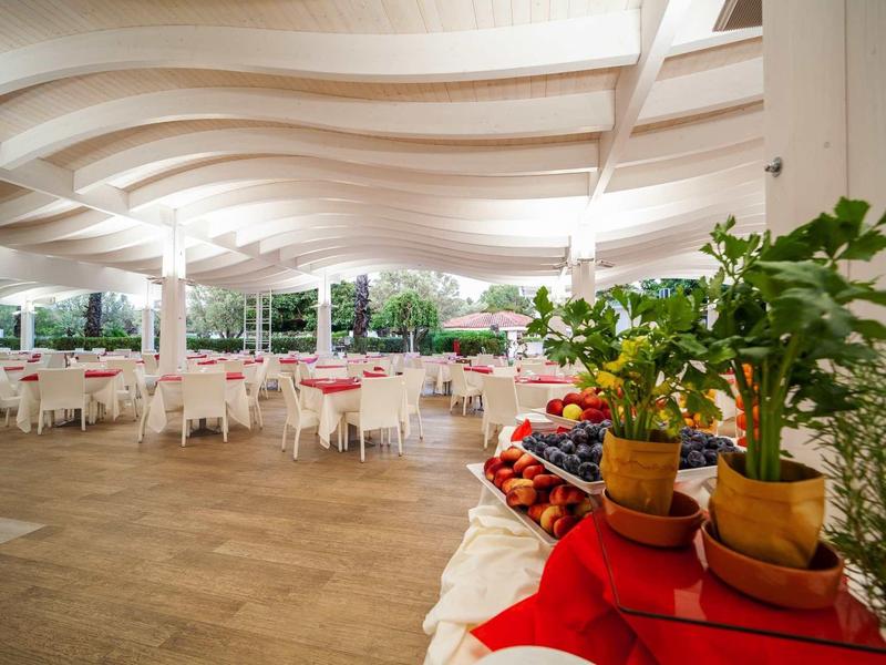 Zadaszony taras z białymi stolikami i krzesłami w hotelu, z roślinami i owocami na pierwszym planie.