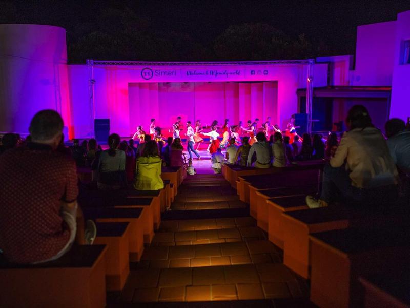 Wieczorny pokaz z publicznością na pomarańczowych ławkach w amfiteatrze na świeżym powietrzu z różowym oświetleniem.