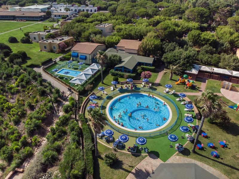 Aerial view of a round pool with umbrellas surrounded by green trees and buildings.