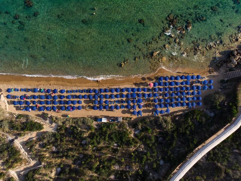 Aerial view of a beach with blue umbrellas and clear water next to a wooded area.