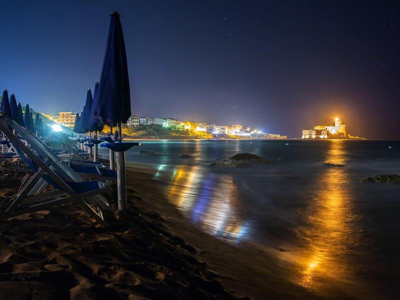 Night beach with lounge chairs, closed umbrellas, and illuminated coastal town in the background.