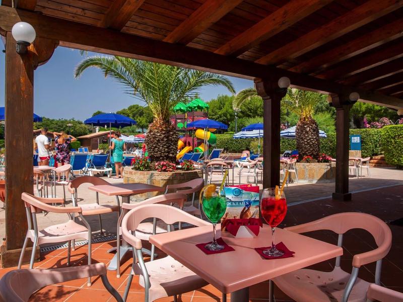 Covered terrace with tables and colorful drinks, in the background pool with palm trees and sun umbrellas
