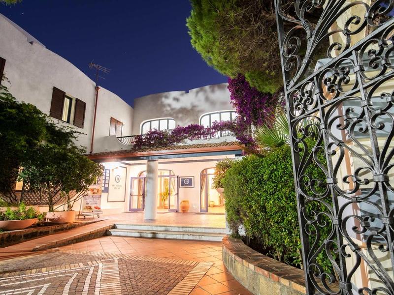 Illuminated hotel garden with terrace, plants, and ornate wrought iron gate at night.