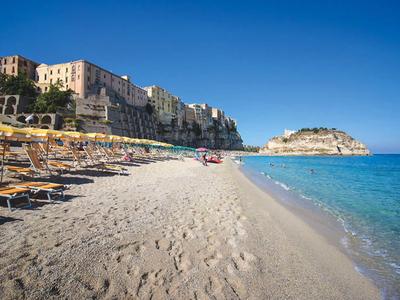 Plage de sable avec chaises longues et parasols devant des bâtiments au bord de la mer sous un ciel clair.