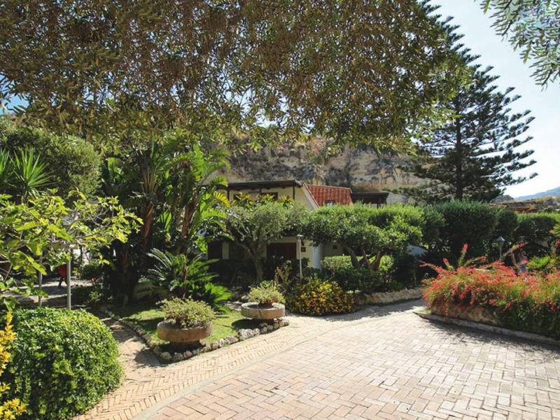 Grand jardin avec des plantes et un chemin en gravier devant un bâtiment hôtelier sous un ciel bleu.