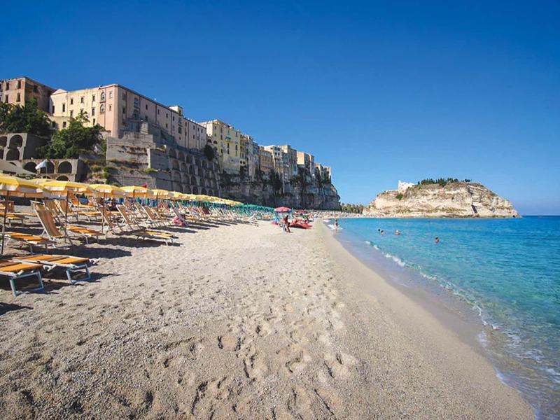 Plage de sable avec chaises longues et parasols devant des bâtiments au bord de la mer sous un ciel clair.