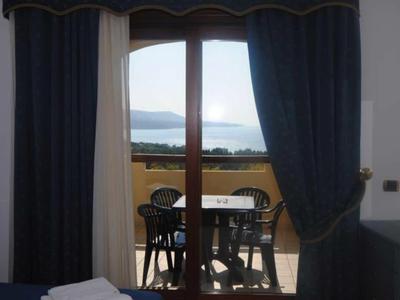 Výhled skrz otevřené závěsy na terasu se stolem a židlemi, za nimi moře a hory.