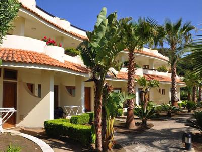 Hotel s bílými budovami, červenými střechami a tropickými rostlinami v zahradě pod jasnou oblohou.