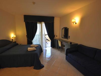 Hotelový pokoj s manželskou postelí, pohovkou, psacím stolem a přístupem na balkon s záclonami.