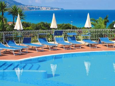 Hotelpool mit blauen Liegestühlen und weißen Sonnenschirmen, Meerblick im Hintergrund.