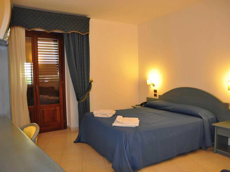 Jednoduchý hotelový pokoj s manželskou postelí, modrým povlečením a dvěma nástěnnými lampami.