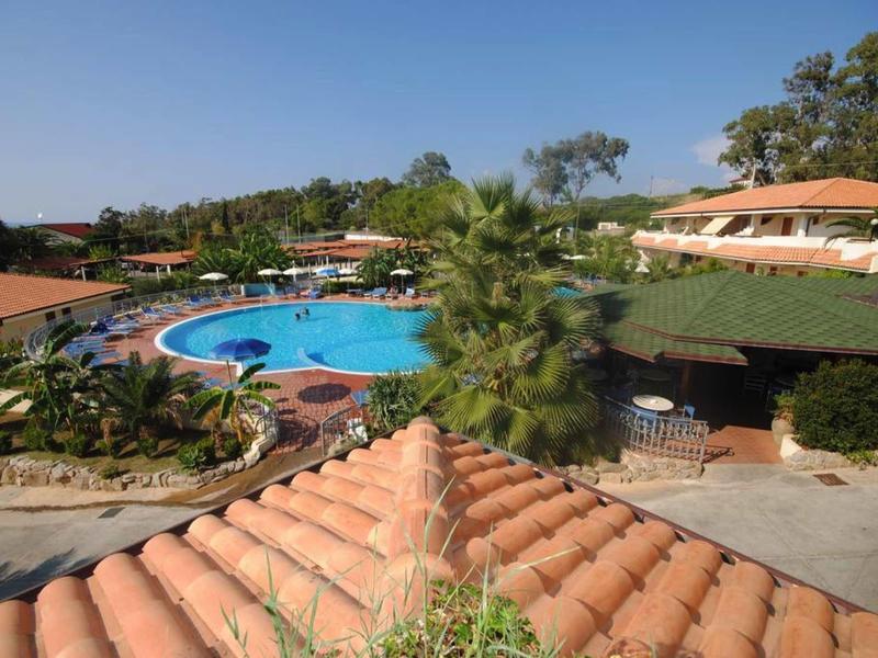 Pohled na hotel s bazénem, palmami a zakřivenými červenými střechami pod jasnou oblohou.