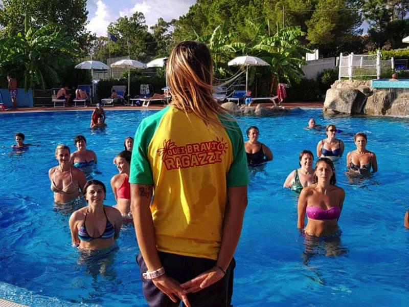 Skupina lidí cvičí vodní aerobik v venkovním bazénu pod vedením instruktora.