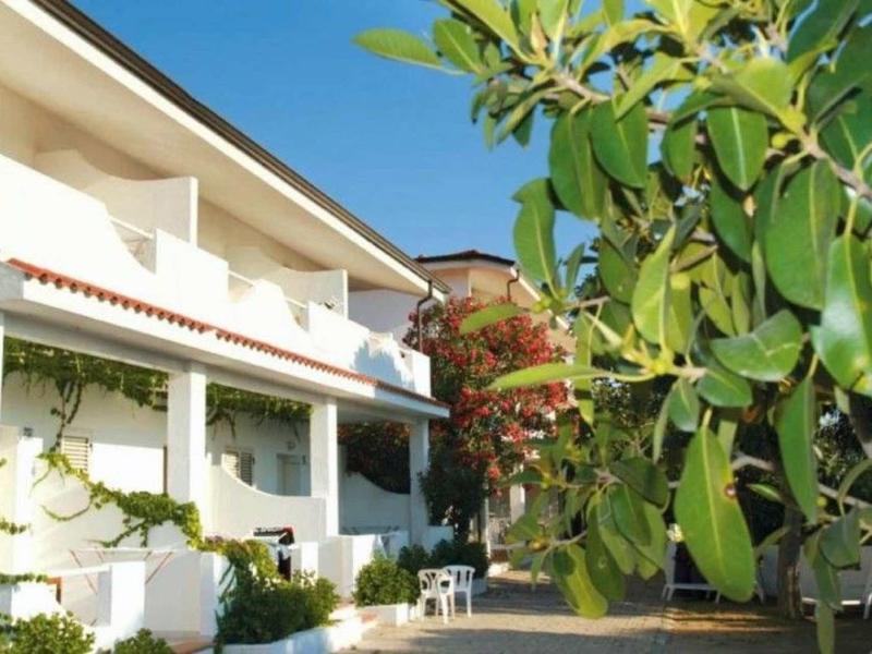 Maison de vacances blanche avec balcon, terrasse et plantes vertes sous un ciel bleu.