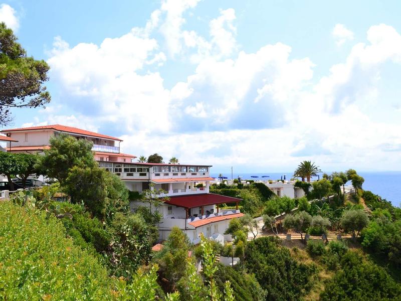 Hôtel sur une falaise verte avec vue sur la mer bleue sous un ciel partiellement nuageux.