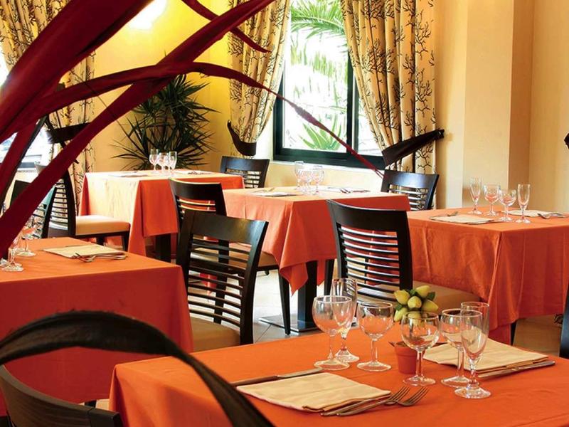 Restaurant mit orange gedeckten Tischen, Holztischen und Stühlen, großen Fenstern und grünen Pflanzen.