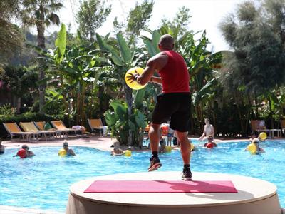 Hombre con ropa deportiva en plataforma dirigiendo aeróbicos acuáticos con participantes en la piscina.