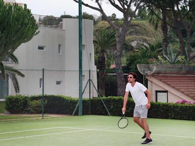 Hombre jugando al tenis en una cancha al aire libre con árboles y edificios al fondo.