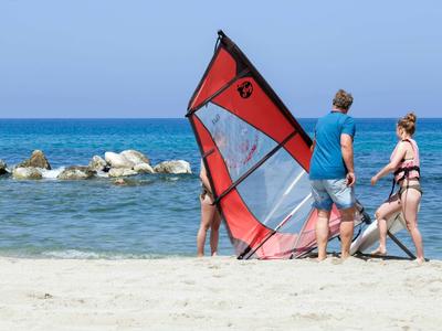 Tres personas en la playa con una vela de windsurf roja y el mar azul al fondo.
