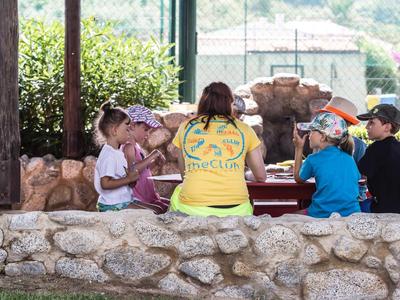 Dzieci siedzą na zewnątrz przy stole z opiekunem na obozie wakacyjnym.
