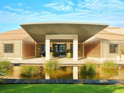 Edificio moderno con grande tettoia, stagno e vegetazione in primo piano sotto cielo azzurro