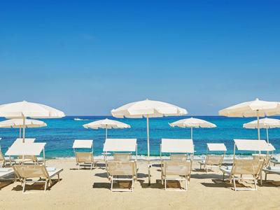 Reihe weißer Liegestühle mit Sonnenschirmen am Sandstrand vor blauem Meer und klarem Himmel.