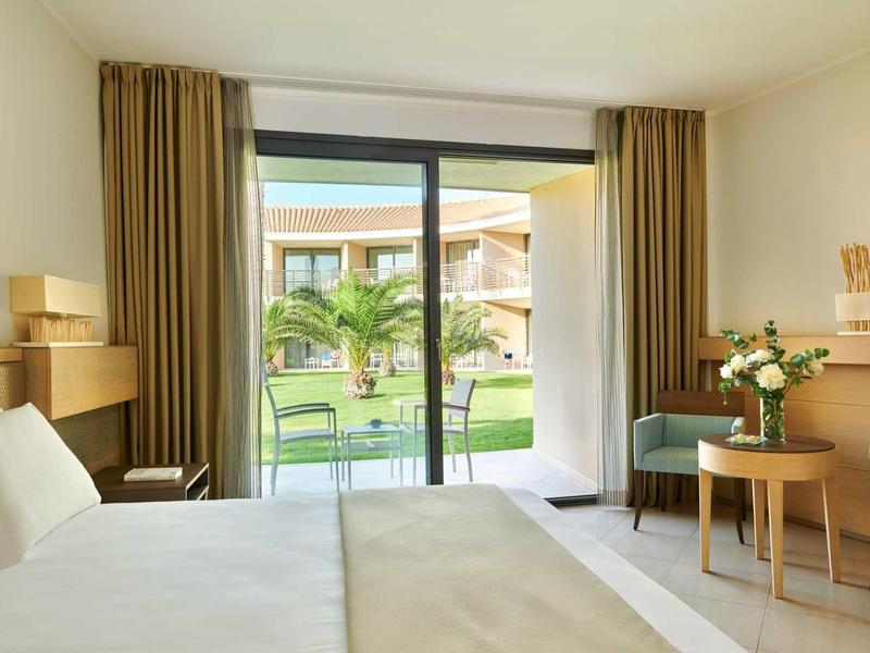 Camera d'hotel luminosa con letto matrimoniale, balcone e vista su giardino con palme.