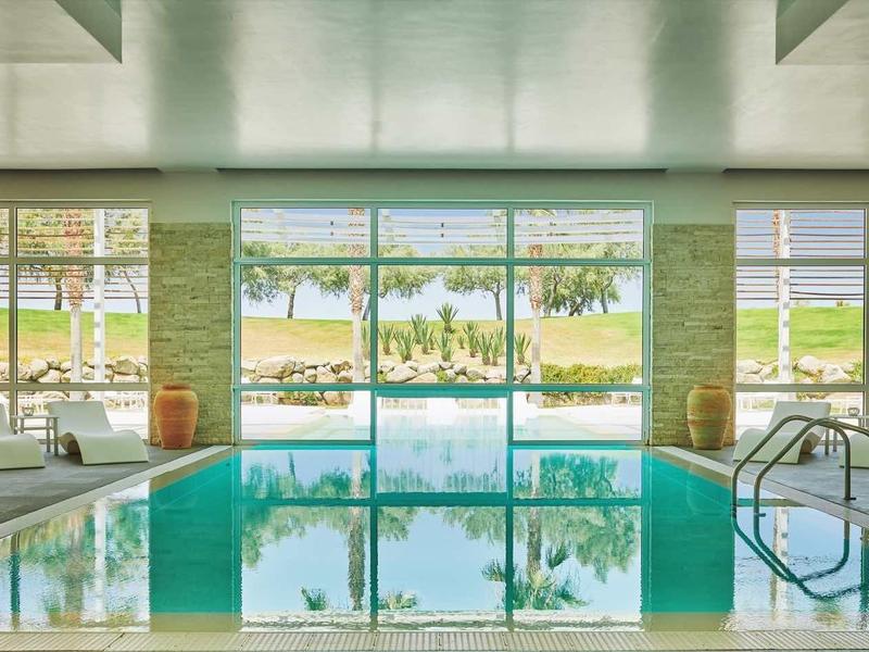 Piscina interna con lettini e grande finestra con vista su giardino e lago.