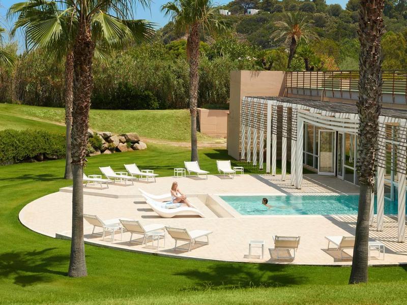 Area piscina con lettini e palme accanto a un edificio moderno, circondata da prato.