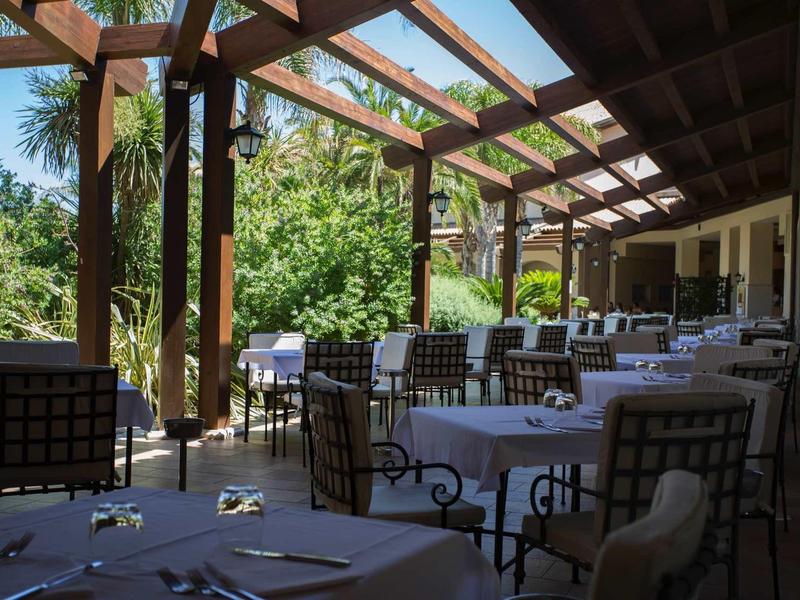 Zadaszony taras na zewnątrz z stołami i krzesłami otoczony zielonymi roślinami.