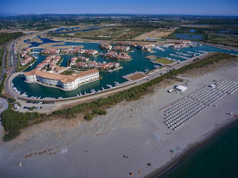 Widok z lotu ptaka nad nadmorskim kurortem z mariną, łodziami i piaszczystą plażą z leżakami.