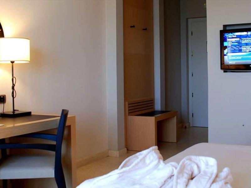 Nowoczesny pokój hotelowy z biurkiem, krzesłem, okrągłym lustrem, telewizorem i łóżkiem z białą pościelą.