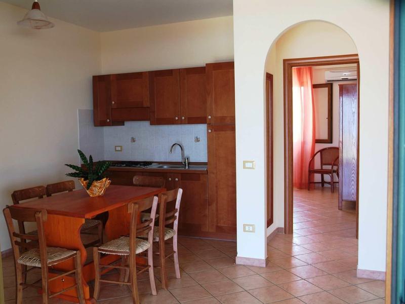 Piccola angolo cottura con mobili in legno e tavolo da pranzo in una stanza luminosa con pavimento piastrellato.