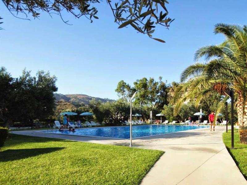 Large hotel pool with sun umbrellas, palm trees, and manicured lawn under blue sky.