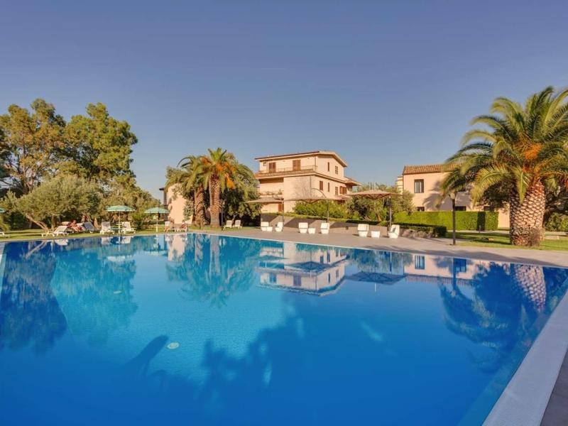 Large blue pool in front of luxury villas with palm trees under clear sky.