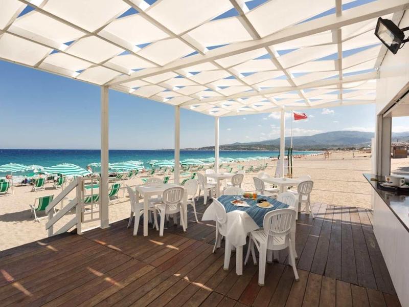 Sunny beach bar with white tables and chairs under canopy overlooking beach and sea.