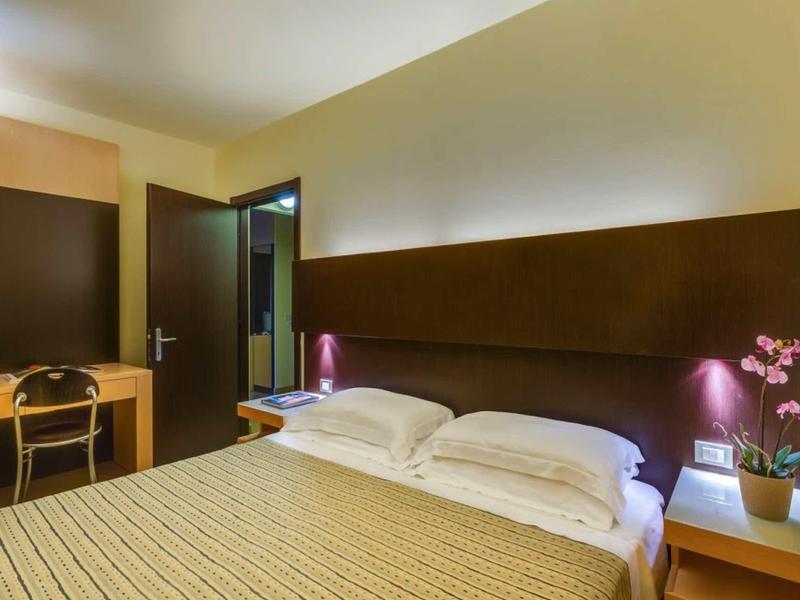 Modern hotel room with double bed, nightstands, chair, and desk in warm tones.