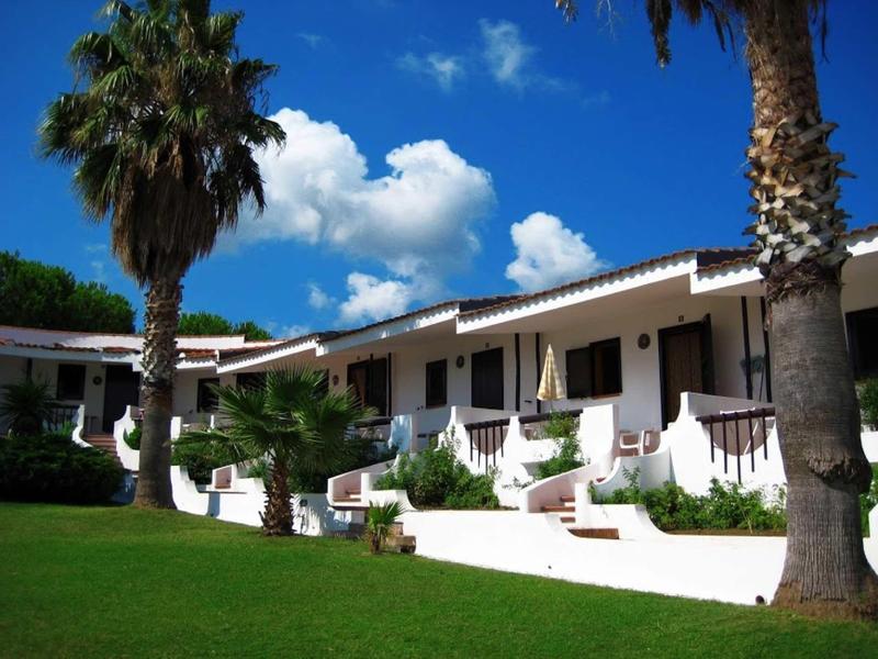 Bílé bungalovy s palmami a zeleným trávníkem pod modrou oblohou v rekreačním středisku.