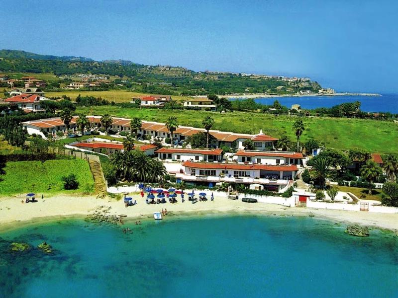 Poklidná pláž s čistou vodou a rekreačními domy v zelené přímořské krajině.