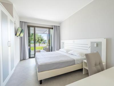 Habitación de hotel luminosa con cama doble, ropa de cama gris, silla y vista a terraza con vegetación.