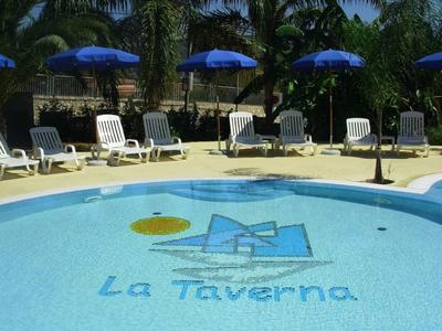 Piscina blu con mosaico di vela e lettini bianchi sotto ombrelloni blu.