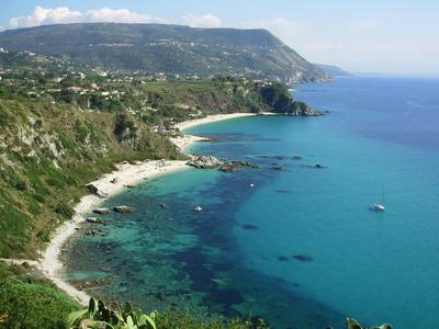 Paesaggio costiero con acqua limpida blu, spiaggia rocciosa e dolci colline sullo sfondo.