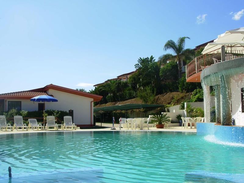 Grande piscina dell'hotel con sdraio, ombrellone e cascata sotto un cielo azzurro.
