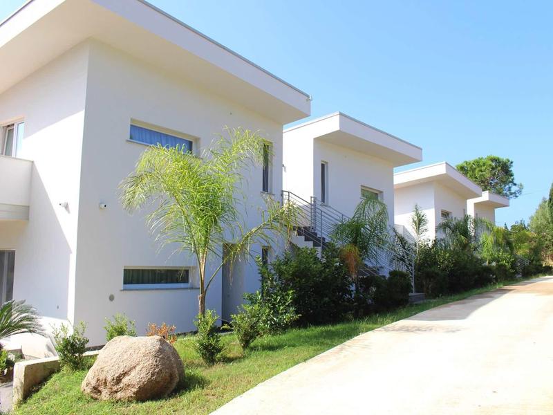 Fila di moderne case vacanza bianche con giardini e palme sotto un cielo terso.