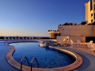 Schwimmbad mit geschwungener Form, umgeben von weißen Liegestühlen vor klarem blauem Himmel.